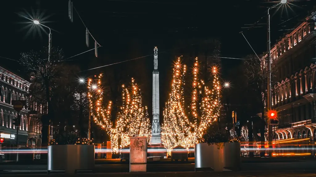 Brīvības piemineklis Rīgā nakts apgaismojumā ar izgaismotiem kokiem un ielas satiksmi.