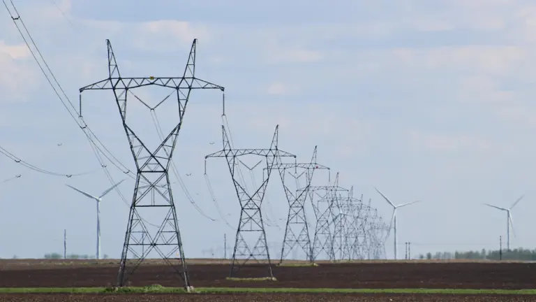 Elektrības pārvades torņi un vēja ģeneratori uz plaša lauka Latvijas ainavā.