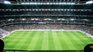 Futbola stadions ar skatītāju pilnām tribīnēm gaidot svarīgu spēli vakara gaismā.