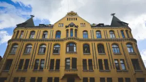 Klasiska Latvijas lauku māja ar koka fasādi un daudziem logiem vasaras dienā.