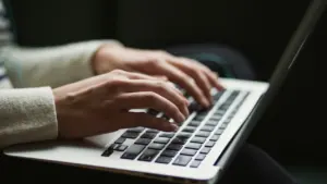 Cilvēka rokas raksta uz sudrabotas klēpjdatora tastatūras tumšā, neitrālā fona vidē.