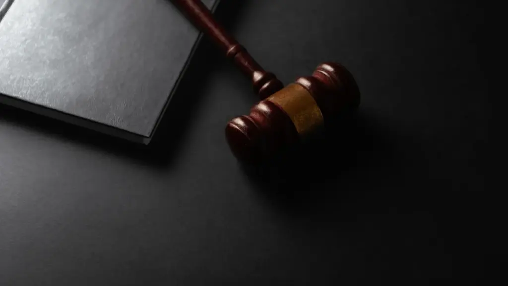 Koka tiesneša āmurs un melna grāmata novietoti uz tumšas, vienkrāsainas virsmas tiesas zāles kontekstā.