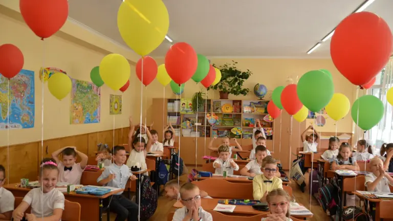 Klasē sēdoši skolēni svin Zinību dienu ar krāsainiem baloniem pie saviem rakstāmgaldiem.