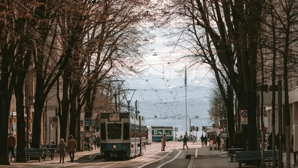 Cilvēki staigā pa Bahnhofstrasse ielu Cīrihē, kurā redzams tramvajs un koki.