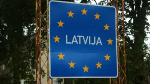 Zila ceļazīme ar uzrakstu Latvija uz valsts robežas starp kokiem vasaras laikā